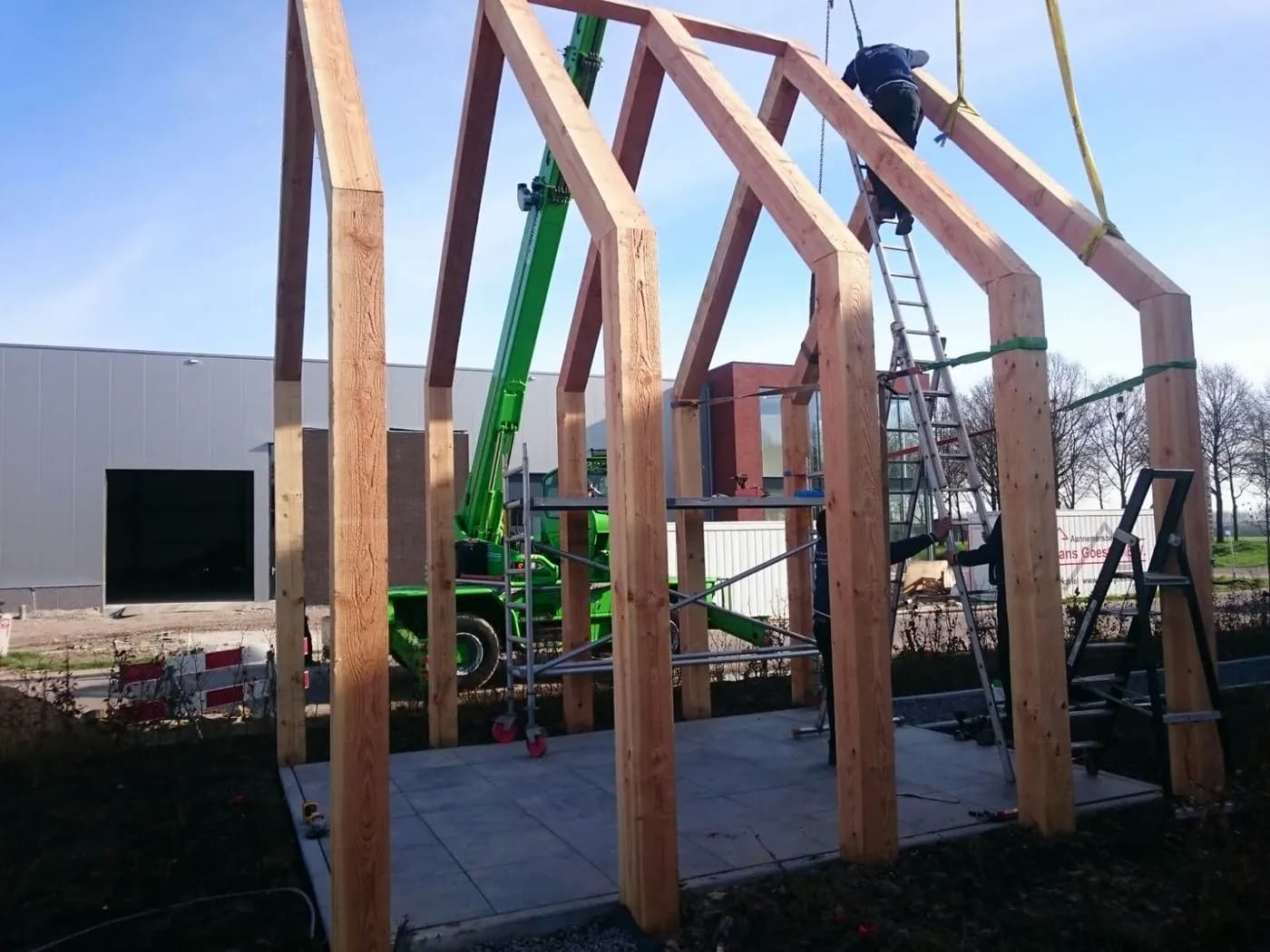 Een kraan zet een houten spantconstructie overeind op een bouwplaats, met een monteur op een ladder ernaast
