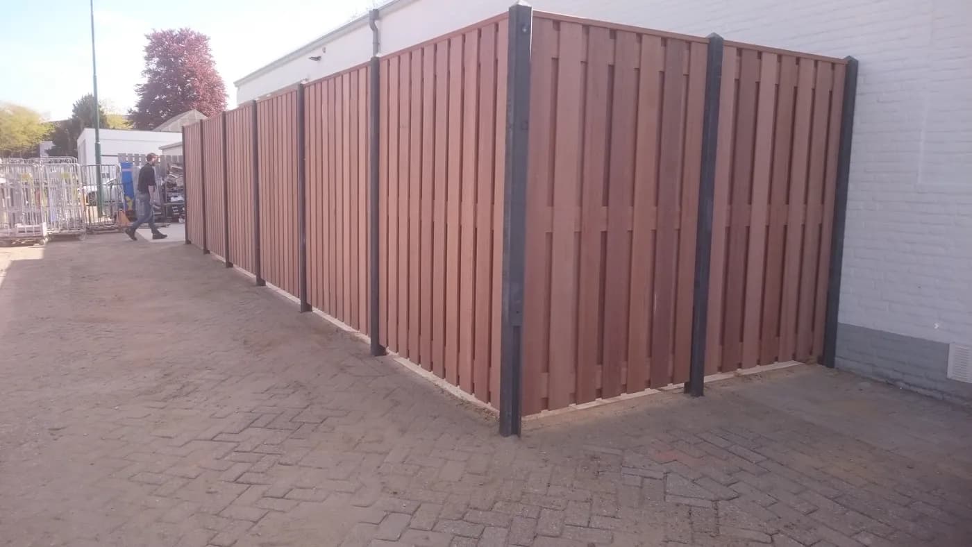 Lange houten schutting met stalen palen, langs een bestrate oprit naast een woning