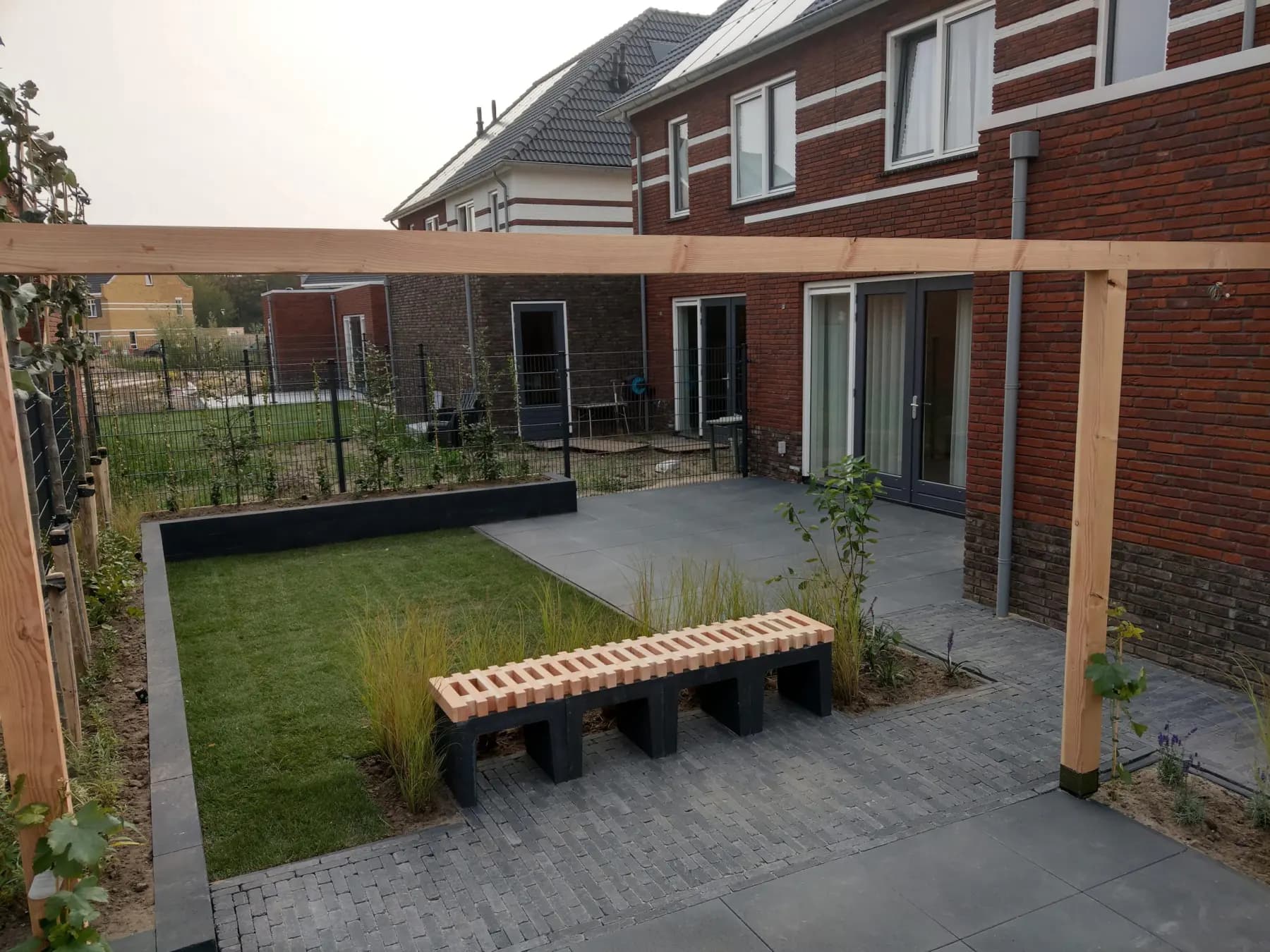 Stadstuin met een houten pergola over het terras, een gemetselde bank, een gazon en verhoogde borders langs de erfgrens