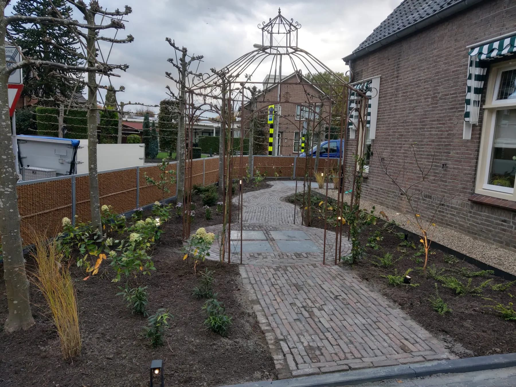 Rustieke tuin bij avond met een klinkerpad, stalen rozenbogen en een koepelprieel, langs borders met jonge beplanting