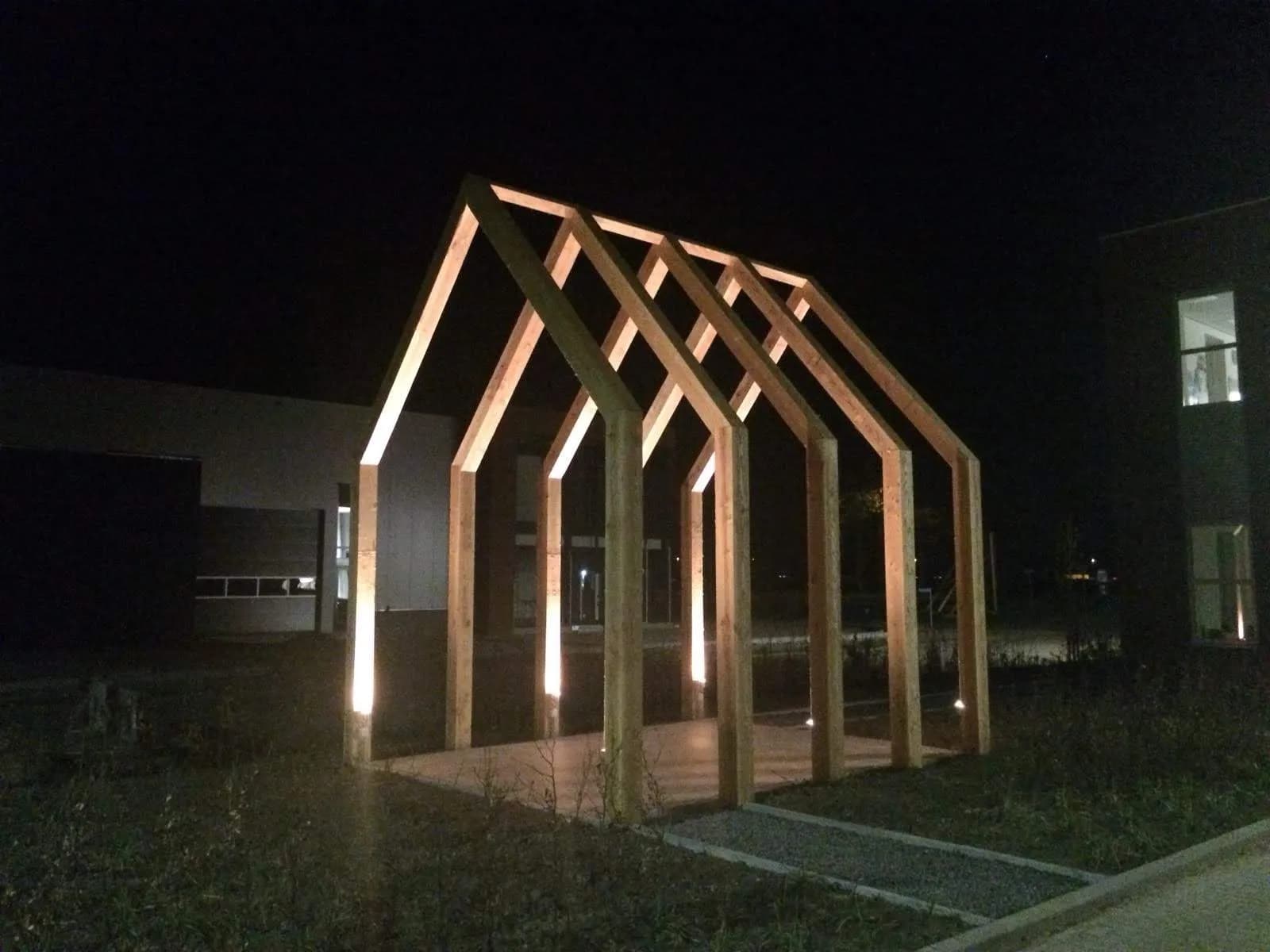 Houten spantconstructie van een overkapping, bij avond uitgelicht op een gazon naast een gebouw