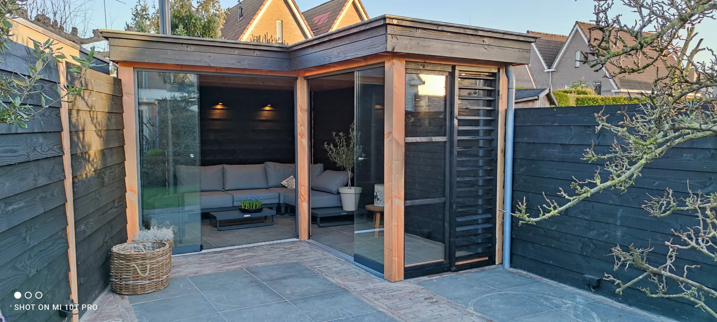 Moderne houten overkapping met een loungehoek eronder, tegen een donkere houten wand en een terras van grote tegels