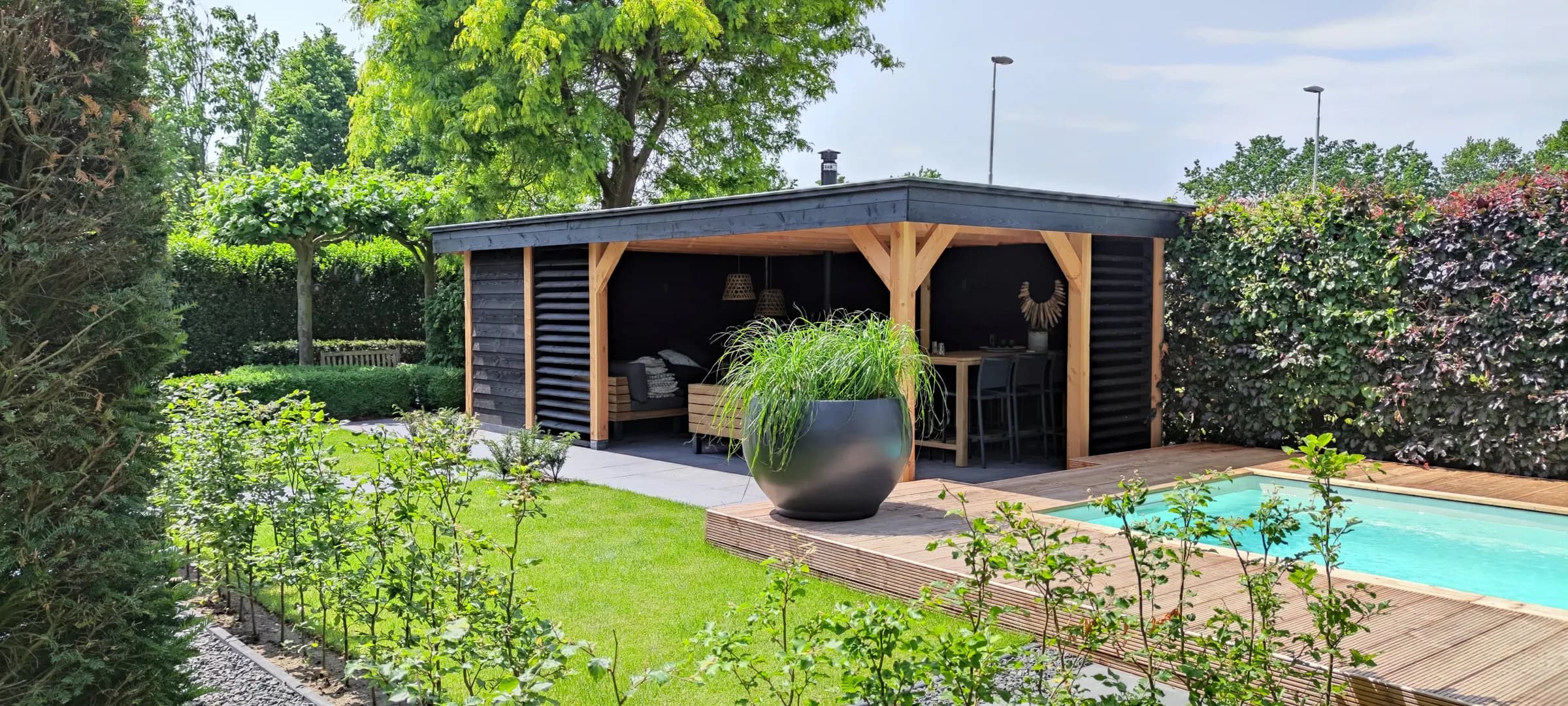 Tuin met een donker afgewerkte overkapping en zithoek, een houten vlonder langs een zwembad en een strak gazon