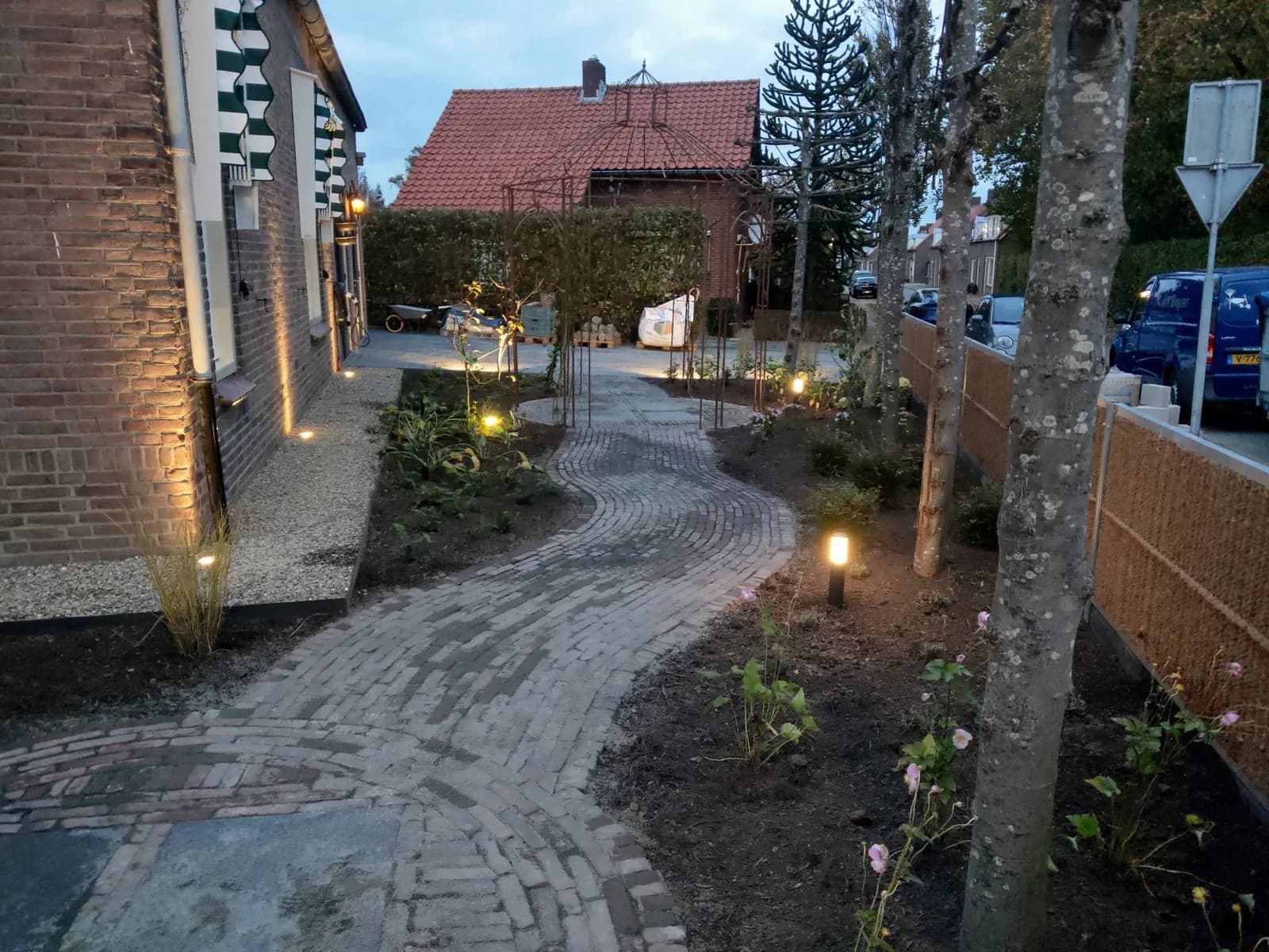 Rustieke tuin bij avond, met verlichte klinkerpaden, jonge bomen en borders