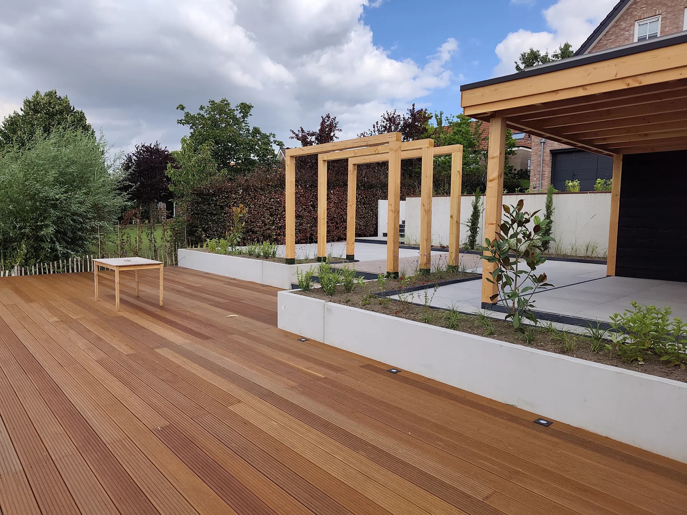 Hardhouten vlonder langs betonnen plantenbakken, met houten pergolaportalen en een overkapping op de achtergrond