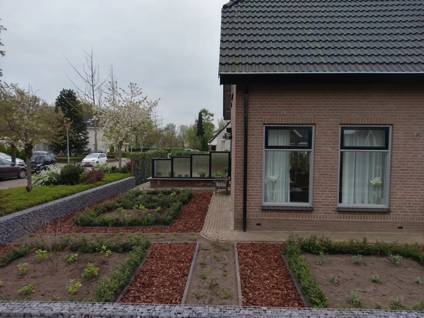 Voortuin met een lage schanskorf van robuuste stenen, vakken met Ilex haag en rode boomschors langs een gemetselde woning